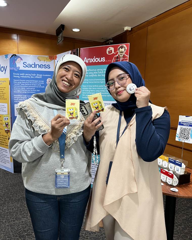 Dua peserta mengangkat kartu bertuliskan Joy dan lencana emosi, di depan panel tentang Sadness dan Anxious.