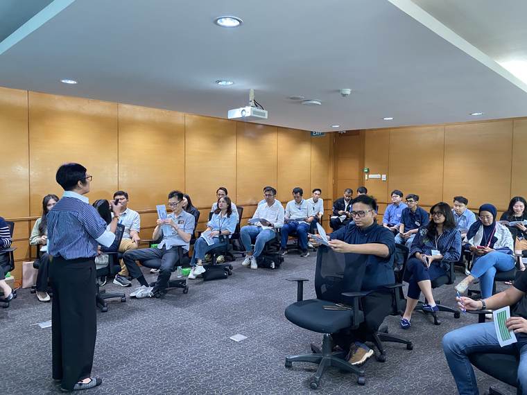 Fasilitator berdiri di tengah lingkaran peserta yang duduk sambil memegang kartu.